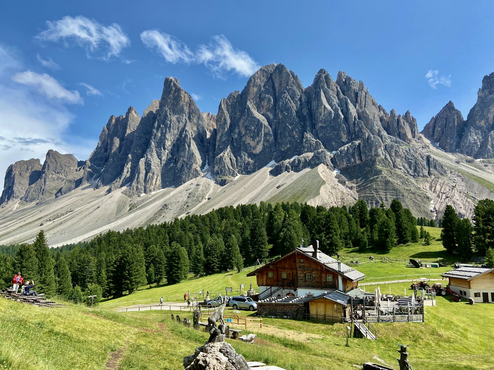 Le punte delle Odle sopra il bosco, con una baita sul prato e il cielo azzurro